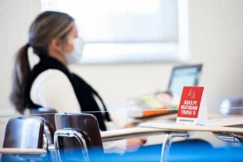 A student sits at her desk on her laptop in the background. In the foreground is a sign that reads "please maintain 6 feet" resting on a desk that is roped off.